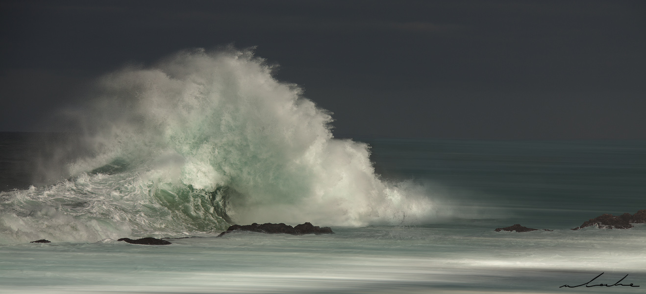 A colour photograph of a wave breaking against some rocks making a fan-like formation